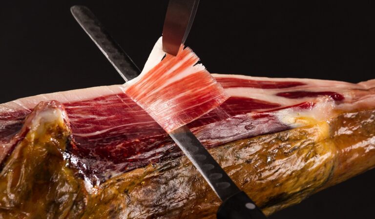 Tast de Pernils 16 Corte de jamón ibérico con dos cuchillos levantando loncha translúcida sobre fondo negro